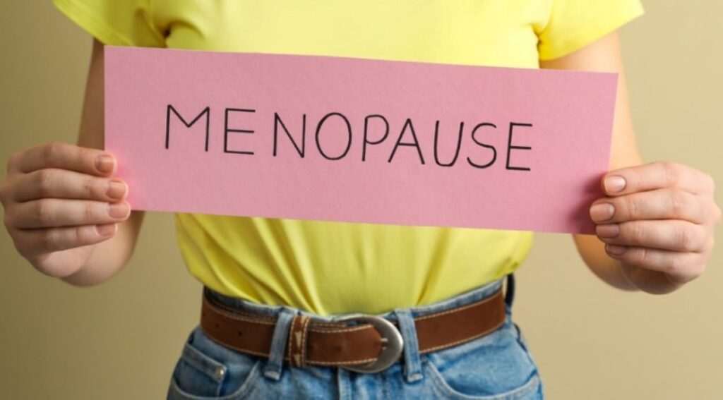 A woman in a yellow tee tucked in tightly holding a cardboard cutout with the word menopause written on it